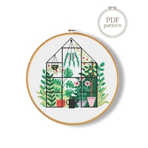 May include: Cross-stitch pattern of a greenhouse filled with plants, in a wooden embroidery hoop. The design features a black frame, green foliage, and pink flowers. The text "PDF pattern" is in a circle at the top.