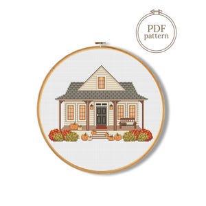Puede incluir: Un patrón de punto de cruz en un bastidor de madera que muestra una casa con porche, calabazas y arbustos. El diseño incluye una puerta oscura, varias ventanas y un tejado. Un gráfico circular en la esquina contiene el texto PDF pattern.