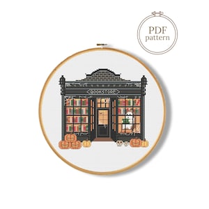 Peut inclure: Un modèle de point de croix dans un tambour à broder en bois représentant une devanture de librairie noire avec des étagères de livres colorés, un fantôme et des citrouilles orange. Le motif inclut le texte BOOKSTORE et une étiquette PDF.
