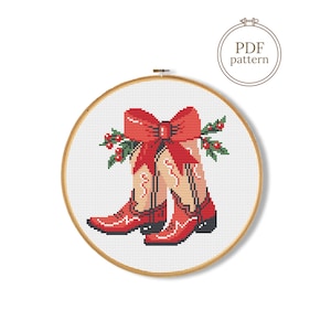 May include: A cross-stitch pattern in a wooden embroidery hoop featuring a pair of red cowboy boots adorned with a large red bow and green holly sprigs. The design is set against a white grid background, with the text PDF pattern in a circle.