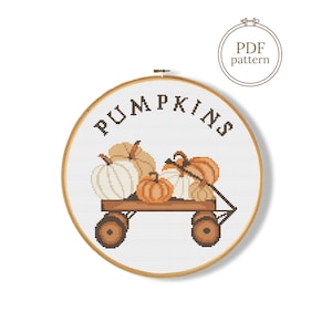 Op de afbeelding: Een kruissteekpatroon in een ronde houten borduurring met een bruine wagen gevuld met witte en oranje pompoenen. Het woord PUMPKINS is in bruine letters boven de wagen geborduurd. Een rond icoon in de hoek bevat de tekst PDF pattern.