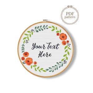Op de afbeelding: Een kruissteekontwerp in een houten borduurring. Het ontwerp toont een bloemenkrans met oranje bloemen en groene bladeren, rondom de tekst "Your Text Here". Een PDF-patroonpictogram staat in de rechterbovenhoek.