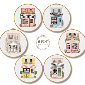 Op de afbeelding: Zes ronde borduurringen met kruissteekpatronen van diverse winkelpuien, waaronder een bakkerij, theehuis en gelateria. Elk ontwerp bevat gedetailleerde architecturale elementen, kleurrijke luifels en planten. De tekst in het midden luidt 6 PDF patterns.