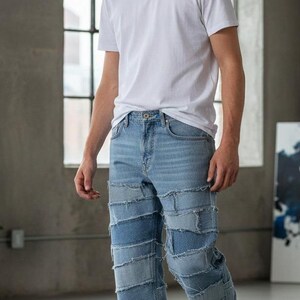 Può includere: Jeans di denim blu chiaro con un design patchwork. I jeans hanno una vestibilità dritta e sono abbinati a una t-shirt bianca e scarpe da ginnastica bianche. Il modello indossa una catena d'argento.