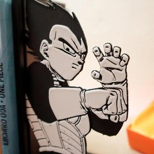 May include: A black and white Vegeta bookend, depicting the character in an action pose. The bookend is positioned beside three "One Piece" manga books. The bookend is made of a solid material and is designed to keep books upright.