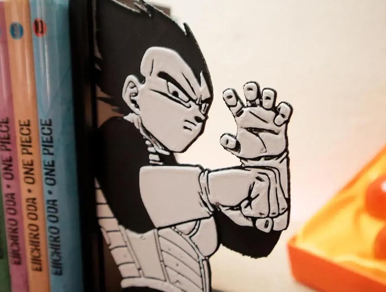 May include: A black and white Vegeta bookend, depicting the character in an action pose. The bookend is positioned beside three "One Piece" manga books. The bookend is made of a solid material and is designed to keep books upright.