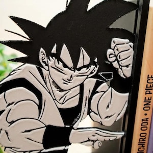May include: A black and white Goku figure, a Dragon Ball character, with a determined expression. The figure is positioned next to a stack of colourful books with the text "ONE PIECE" on the spines. The books are numbered 5 and 6.