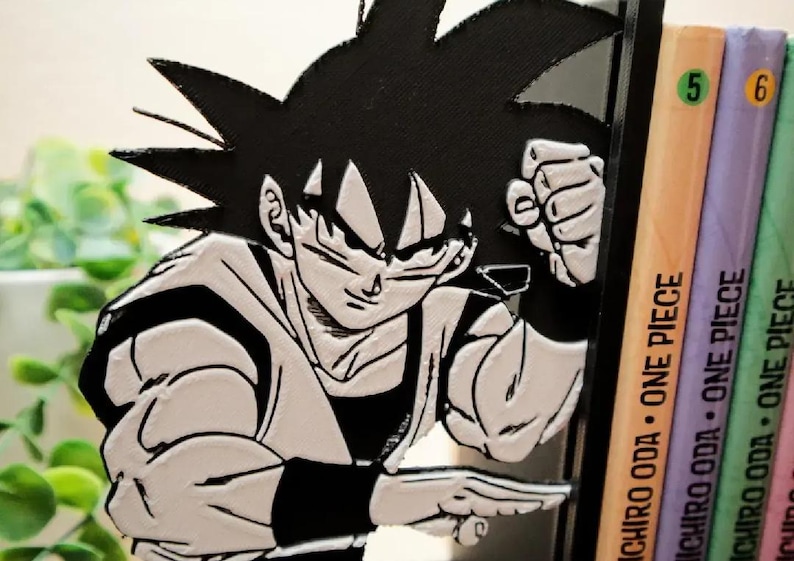 May include: A black and white Goku figure, a Dragon Ball character, with a determined expression. The figure is positioned next to a stack of colourful books with the text "ONE PIECE" on the spines. The books are numbered 5 and 6.