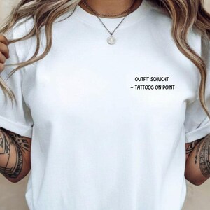 Könnte beinhalten: Weißes T-Shirt mit dem Text "Outfit Schlicht - Tattoos on Point" in Schwarz. Das T-Shirt hat einen Rundhalsausschnitt und wird mit einer blauen Jeans getragen. Die Person, die das T-Shirt trägt, hat mehrere Tätowierungen an Armen und Handgelenken.
