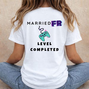 Könnte beinhalten: Ein weißes T-Shirt mit einem Aufdruck auf der Rückseite, der den Text MARRIED AF zeigt, wobei AF mit einem Glitch-Effekt stilisiert ist. Unter dem Text befindet sich eine türkisfarbene Videospiel-Controller-Grafik mit Kabel und LEVEL COMPLETED.