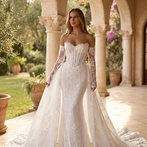 Könnte beinhalten: Ein weißes Brautkleid mit schulterfreiem Design, langen Spitzenärmeln und einem figurbetonten Oberteil. Das Kleid hat detaillierte florale Spitze und eine fließende Schleppe. Die Braut befindet sich in einer Außenkulisse mit Steinsäulen und Bögen.