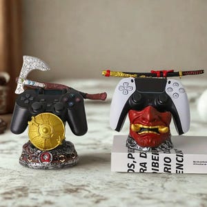 May include: Two decorative controller stands. One features a black controller with a silver axe and a gold disc. The other has a white and black controller resting on a red demon mask, with a miniature sword on top.