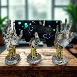 Könnte beinhalten: Ein schwarzer Videospiel-Controller wird von zwei silber- und goldfarbenen Roboterhand-förmigen Ständern gehalten. Der Controller hat leuchtende blaue und violette Akzente. Die Ständer stehen auf einer Holzoberfläche.