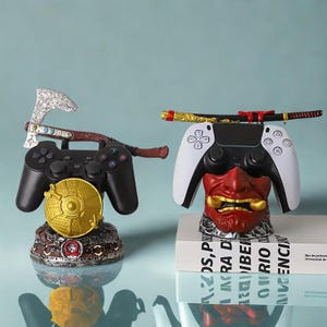 Könnte beinhalten: Zwei Gamecontroller-Ständer. Einer zeigt einen schwarzen Controller mit einer silbernen Axt und einer goldenen Scheibe. Der andere hat einen weiß-schwarzen Controller mit einer roten Dämonenmaske und einem Katana-Schwert. Beide sind dekorativ.