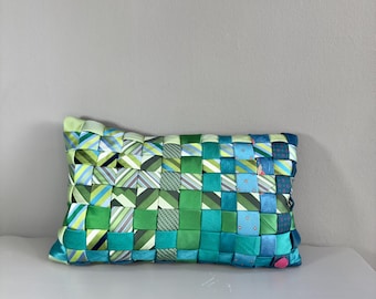 Op de afbeelding: Een rechthoekig kussen met een geweven ontwerp. Het kussen heeft een patchwork van groene, blauwe en blauwgroene linten. Sommige linten hebben strepen of stippen. Het kussen ligt op een wit oppervlak.