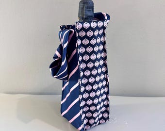 Op de afbeelding: Een marineblauwe wijnfleszak met een roze en wit patroon. De tas is gemaakt van hergebruikte stropdassen. Eén kant heeft roze strepen, terwijl de andere een herhalend bloemenpatroon heeft. De tas is ontworpen om een standaard wijnfles te bevatten.