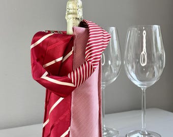 Op de afbeelding: Een rood-wit gestreepte wijnfleszak met een champagnefles erin. De tas heeft een handvat van rode stof met witte diagonale strepen. Twee wijnglazen staan op de achtergrond.