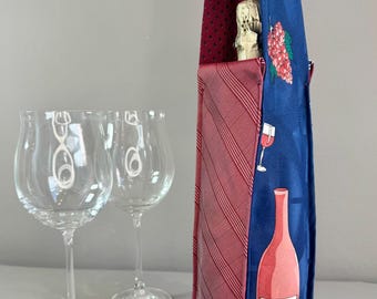 Op de afbeelding: Een wijntas gemaakt van hergebruikte stropdassen. De tas heeft een rood-blauwe kleurstelling met wijn-thema afbeeldingen. Twee heldere wijnglazen staan op de voorgrond en een fles champagne zit in de tas.
