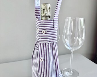 Op de afbeelding: Een wijnflesdrager gemaakt van een gestreept overhemd, met een handvat met de tekst "PARADISENKNITS". De drager heeft knopen en bevat een champagnefles. Een wijnglas staat ernaast.