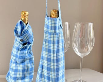 Op de afbeelding: Twee wijnfleshoezen van blauw-wit geruite stof. Eén fles is verpakt en de andere zit in een houder met een handvat. Twee wijnglazen staan op de achtergrond. De flessenhoezen zijn een unieke manier om wijn cadeau te geven.