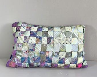 May include: Rectangular decorative pillow woven from repurposed neckties. The pillow features a patchwork design with various colors, including purple, green, and gray. The pillow's dimensions are approximately 12 inches long and 8 inches wide.