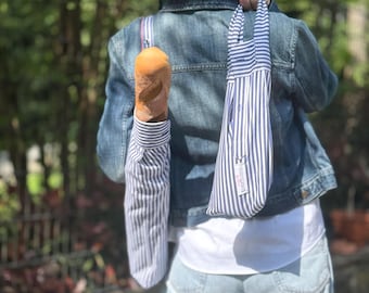 Op de afbeelding: Een persoon draagt een vers stokbrood in een blauw-wit gestreepte broodzak. De tas is gemaakt van een overhemd en heeft een bijpassende draagtas. De persoon draagt een spijkerjasje en een lichtblauwe jeans.