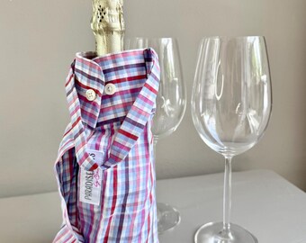 Op de afbeelding: Een champagnefles is gekleed in een geruite shirtvormige wijnfleshoes in rood, wit en blauw. De hoes heeft knopen en een klein zakje. Twee wijnglazen staan achter de fles.