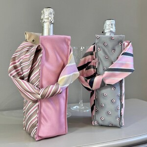May include: Two champagne bottle covers, one pink and one gray, each adorned with a necktie. The pink cover has a striped tie, while the gray cover features a diamond pattern. The ties are wrapped around the bottles, creating a decorative effect.