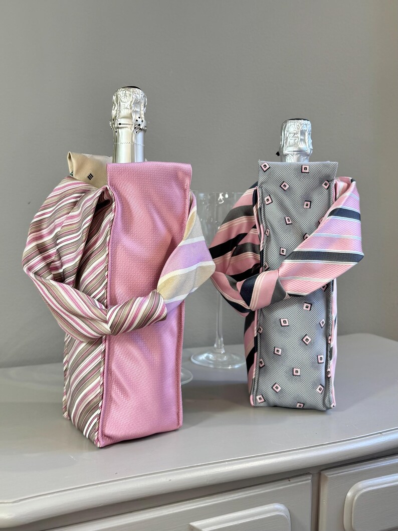 May include: Two champagne bottle covers, one pink and one gray, each adorned with a necktie. The pink cover has a striped tie, while the gray cover features a diamond pattern. The ties are wrapped around the bottles, creating a decorative effect.