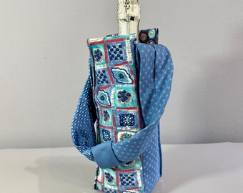 Op de afbeelding: Een blauwe stoffen wijnfleszak met een decoratief patroon van vierkanten in turquoise, rood en marineblauw. De tas heeft een handvat van blauwe stof met een patroon. De fles is gedeeltelijk zichtbaar.