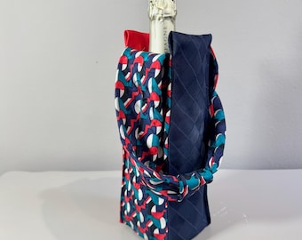 May include: A champagne bottle with a decorative fabric wine bag. The bag features a navy blue, red, and teal geometric pattern on one side and a solid navy blue quilted fabric on the other. A red bow is visible.