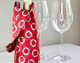 Op de afbeelding: Een rode wijnfleszak met een nautisch reddingsboei patroon. De tas bevat een champagnefles, met twee wijnglazen op de achtergrond. Het reddingsboei ontwerp heeft witte, blauwe en gele accenten.