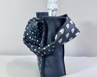 Op de afbeelding: Een zwarte wijnfleszak met een decoratieve strik. De strik is zwart met witte vierkante en hartvormige patronen. De tas is ontworpen om een wijnfles te bevatten en voegt een vleugje elegantie toe.