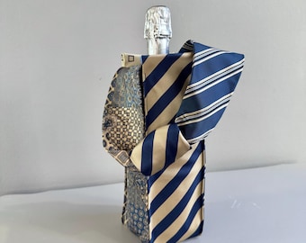 Op de afbeelding: Een champagnefles verpakt in een patchwork van stropdassen met patronen. De stropdassen hebben strepen en geometrische patronen in blauw, beige en crème. De folie bovenop de fles is zichtbaar.