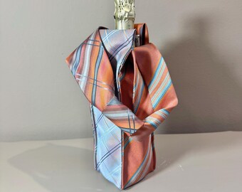 May include: A champagne bottle is wrapped in a decorative fabric made from repurposed neckties. The fabric features a mix of orange, blue, and white stripes and plaid patterns. The bottle's gold foil top is visible.