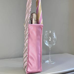 May include: A pink wine bottle carrier with a striped handle. The carrier is holding a champagne bottle. A wine glass is next to the carrier. The handle is made from repurposed neckties with stripes of brown, pink, and white.