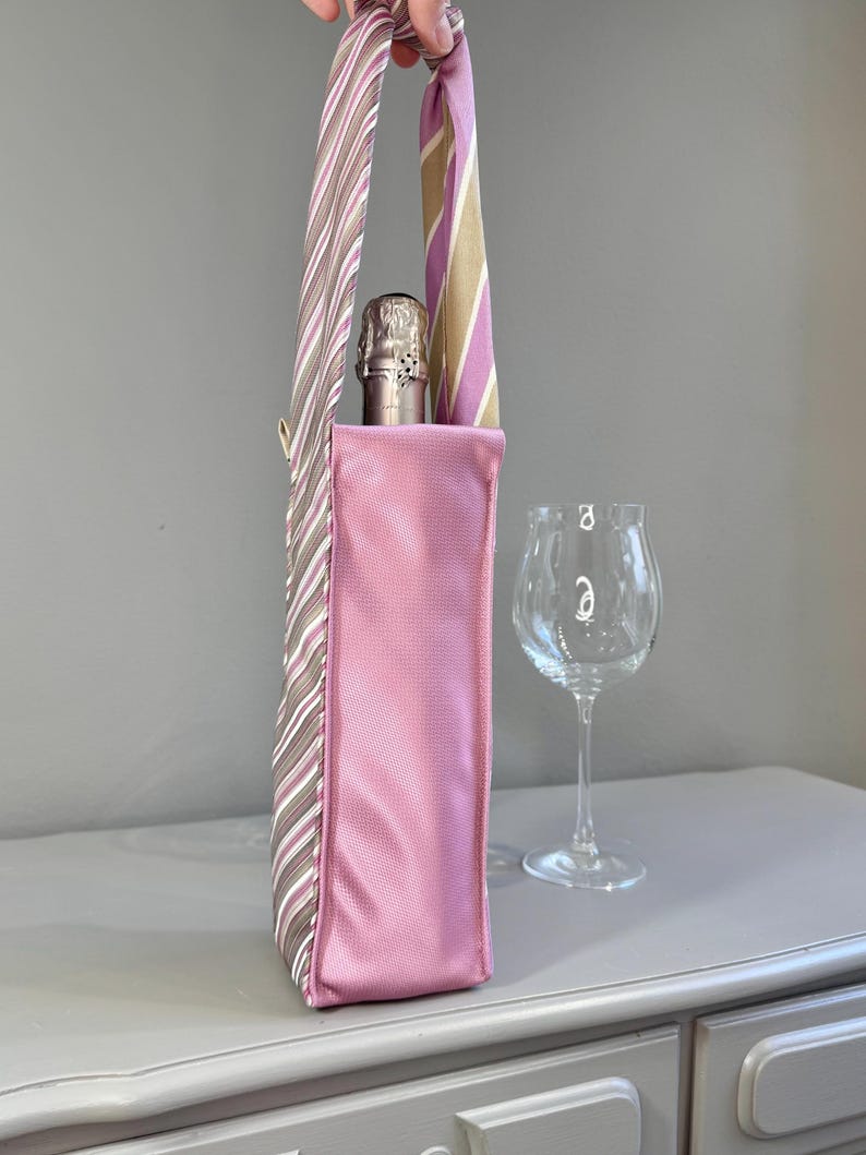 May include: A pink wine bottle carrier with a striped handle. The carrier is holding a champagne bottle. A wine glass is next to the carrier. The handle is made from repurposed neckties with stripes of brown, pink, and white.