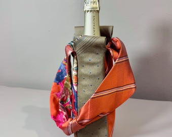 Op de afbeelding: Een champagnefles is versierd met een unieke flesverpakking gemaakt van hergebruikte stropdassen. De verpakking heeft een combinatie van patronen en effen stropdassen in tinten oranje, blauw, roze en grijs, waardoor een stijlvol en milieuvriendelijk accessoire ontstaat.