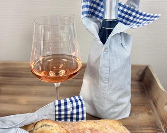 Op de afbeelding: Een champagnefles verpakt in een blauw-wit gestreept overhemd, met een blauw-wit geruite kraag. Een glas rosé wijn en een brood maken de scène compleet, wat duidt op een picknick.