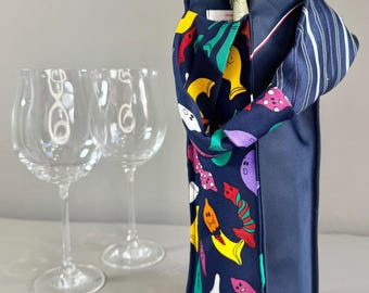 Op de afbeelding: Een marineblauwe wijnfleszak met een kleurrijk patroon van grillige wezens. De tas bevat een champagnefles. Twee heldere wijnglazen staan op de achtergrond. De tas heeft een gestreept stoffen detail.