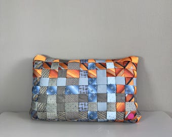 Op de afbeelding: Een rechthoekig kussen geweven van patronen stropdassen in tinten oranje, blauw en grijs. Het kussen heeft een getextureerd oppervlak en een zachte uitstraling, geschikt voor interieurdecoratie.