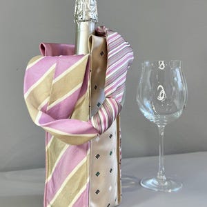 May include: A champagne bottle in a decorative holder made from repurposed neckties. The holder features pink, beige, and striped patterns. A clear wine glass stands beside the bottle, set against a neutral background.