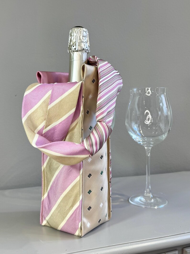May include: A champagne bottle in a decorative holder made from repurposed neckties. The holder features pink, beige, and striped patterns. A clear wine glass stands beside the bottle, set against a neutral background.