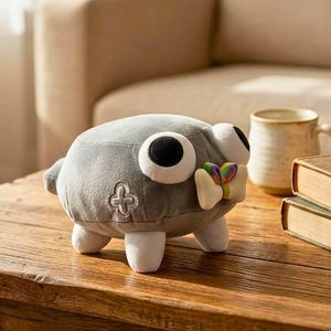May include: A gray plush toy with large, round eyes and a small, colorful bow. The toy has white feet and a four-leaf clover emblem. It is sitting on a wooden table.