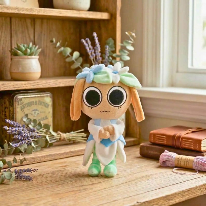 May include: A plush doll with large, round eyes and a floral headpiece. The doll has tan skin, long ears, and is wearing a white and blue outfit with green accents. It is sitting on a wooden surface near a window.