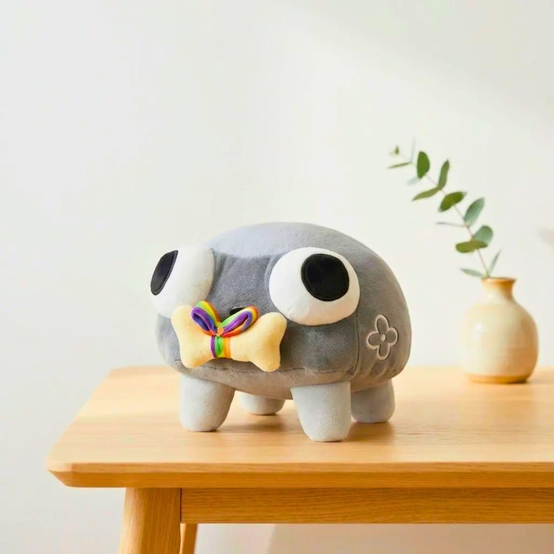 May include: A gray plush toy shaped like a creature with large white eyes and black pupils. It has a yellow bone-shaped accessory with a rainbow ribbon and a small flower emblem. The toy is on a wooden surface.
