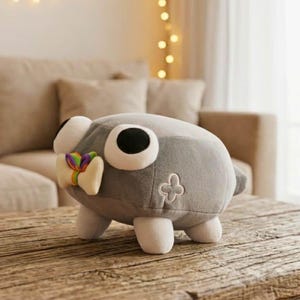 May include: A gray plush toy with large white and black eyes, a colorful bow, and four white feet. The toy has a cross-shaped embroidered detail on its side and is sitting on a wooden table.