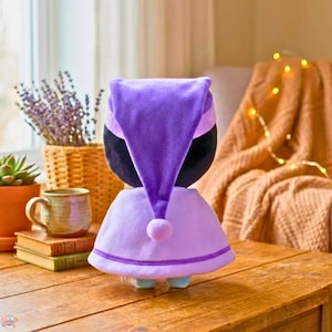 Pu&ograve; includere: Una bambola di peluche con cappuccio e mantello viola. La bambola ha la testa nera e indossa un mantello viola chiaro con finiture viola scuro. Un piccolo pompon viola chiaro &egrave; alla base del cappuccio.