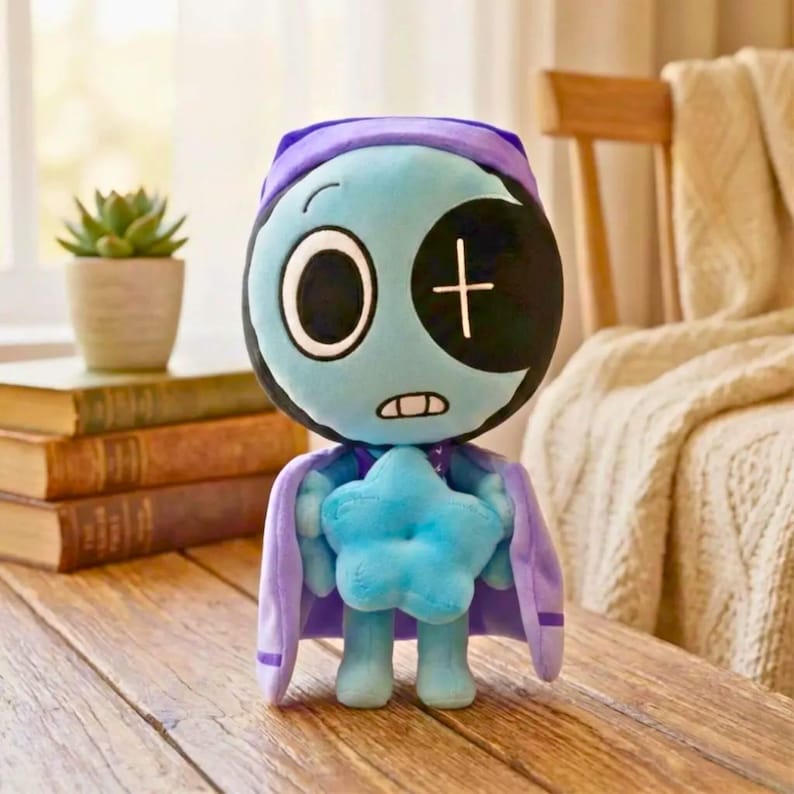 Pu&ograve; includere: Una bambola di peluche con un viso azzurro, un occhio grande e una benda nera con una croce bianca. Indossa un berretto e un mantello viola e tiene in mano una stella blu. La bambola &egrave; su una superficie di legno.