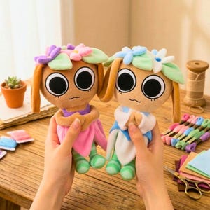 May include: Two plush dolls with large eyes, flower crowns, and long ears. One doll wears a pink outfit, the other a blue outfit. The dolls are held in hands, with craft supplies in the background.